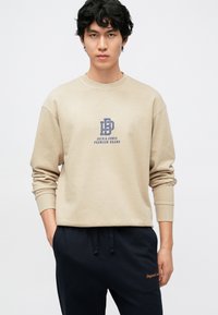 Beige sweatshirt met geribbelde manchetten en een ronde hals, met een marineblauw logo en tekst, gecombineerd met donkerblauwe broek met trekkoorden.