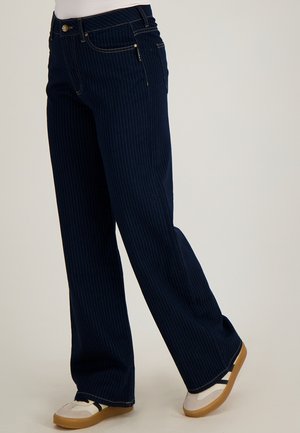 Marineblauwe wijde pijpenbroek met krijtstreep, gedragen met witte sneakers met bruine zolen, getoond van taille tot voeten tegen een effen achtergrond.