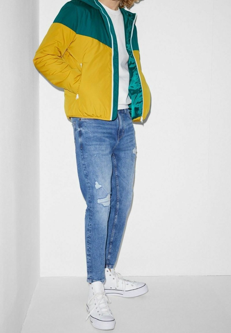 Colorblock-Bomberjacke in Gelb und Türkis, mit Kapuze und Reißverschluss. Wird über einem weißen Hemd getragen und mit verwaschenen blauen Jeans sowie weißen Sneakern kombiniert.