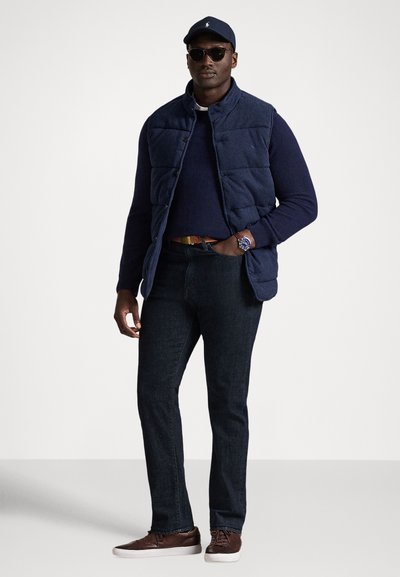 Granatowa pikowana kamizelka na granatowym swetrze, ciemne dżinsy i brązowe buty. Dodatki to czapka i okulary przeciwsłoneczne. Casualowy, warstwowy strój.