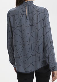 Langærmet blouse i blødt, let stof. Den har en blå baggrund med et geometrisk sort linjemønster og en volangkrave.