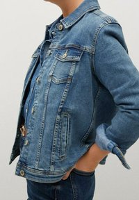 Chaqueta de mezclilla en azul claro con corte clásico, bolsillos frontales, cierre de botones y un ligero desgaste en las mangas y el dobladillo.