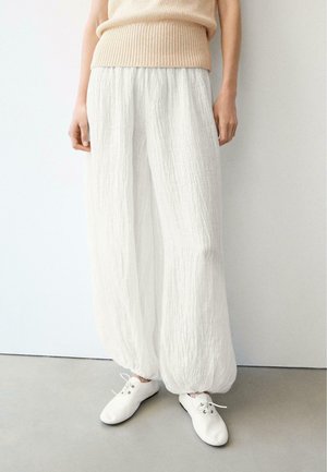 Osoba ubrana w luźne, pogniecione białe spodnie ze ściągaczami przy nogawkach, beżowy dzianinowy top bez rękawów oraz białe sznurowane buty, stojąca przy białej ścianie.