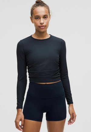 Schwarzes langärmeliges Oberteil mit gerippter Textur, kombiniert mit hoch taillierten schwarzen Shorts. Beide Kleidungsstücke sind figurbetont geschnitten.