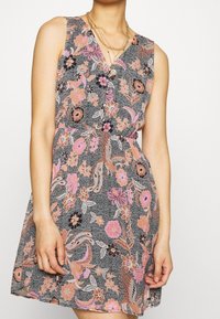 Vestido sin mangas con estampados de flores en rosa, melocotón y negro, llevado con collares de cadenas doradas en capas, en una persona con los brazos relajados a los lados.