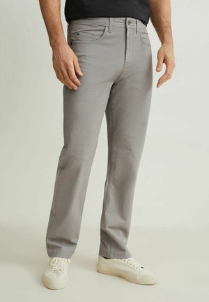 Mężczyzna w jasnoszarych prostych spodniach i białych tenisówkach stoi na tle jednolitego jasnego tła.