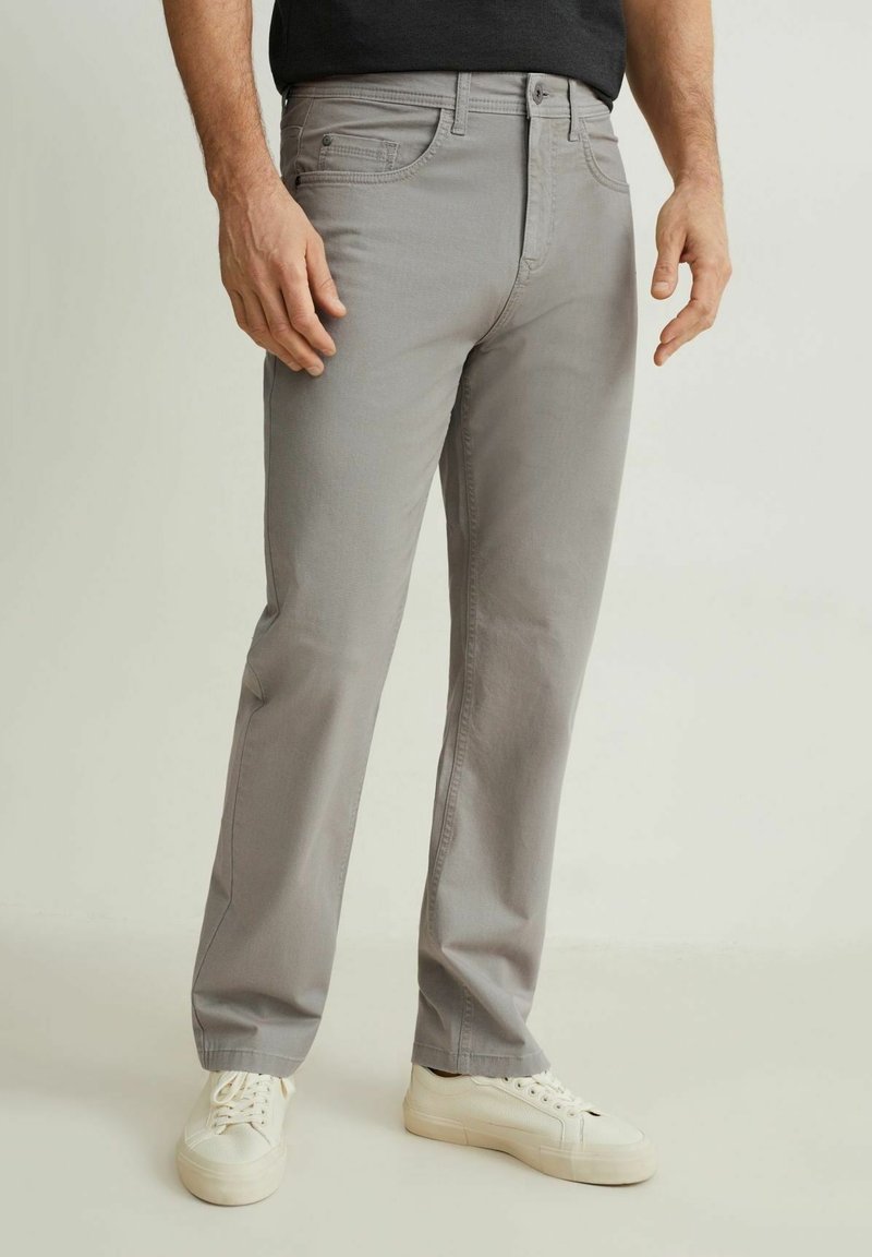 Homme portant un pantalon gris clair droit et des baskets blanches, debout devant un fond clair uni.
