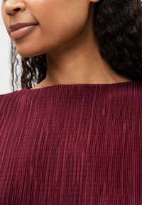 Πλαισιωμένο τοπ σε μπορντώ χρώμα με φαρδιά λαιμόκοψη, υφαντό ύφασμα και κατακόρυφες πτυχώσεις, που αναδεικνύει ομαλή υφή και διακριτική γυαλάδα.