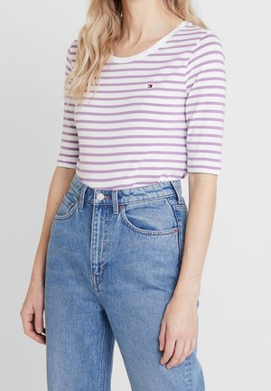 Nainen pukeutuneena laventelin ja valkoisen raidoitettuun kolmivartiseen paitaan, joka on tucking korkeavyötäröisiin sinisiin farkkuihin, seisten yksinkertaisen taustan edessä.