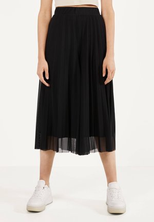 Mujer con una falda midi plisada negra combinada con zapatillas blancas, de pie frente a un fondo claro y liso.