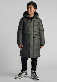 Zeleni puhovka s kapuco, ki ima gumb za zapiranje spredaj, dva sprednja žepa in prešito teksturo. Nosijo jo nad črnimi hlačami in supergami.