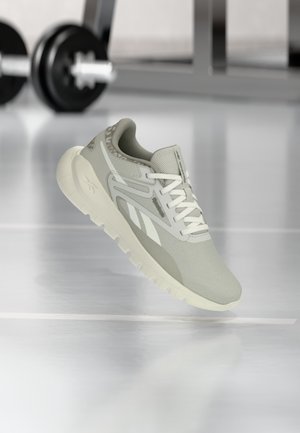 Zapatilla Reebok gris claro con cordones y suela blancos, situada frente a pesas de gimnasio desenfocadas sobre un suelo brillante.