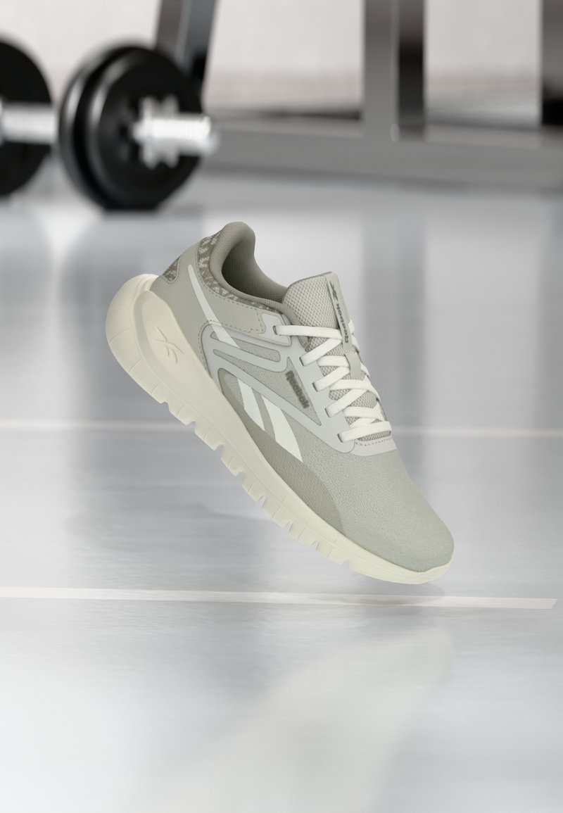 Ανοιχτό γκρι αθλητικό παπούτσι Reebok με λευκά κορδόνια και σόλα, τοποθετημένο μπροστά σε θολά βάρη γυμναστηρίου πάνω σε γυαλιστερό πάτωμα.