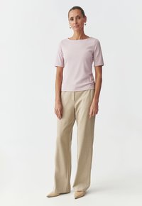 Top rosa chiaro a maniche corte abbinato a pantaloni beige a gamba larga e scarpe beige con punta, su sfondo bianco semplice.