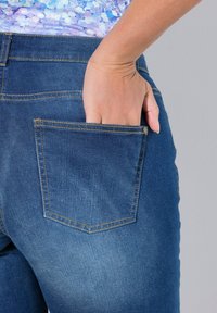 Tmavě modré džíny z denimu se zadní kapsou, ve které je částečně vložená ruka. Mají zlaté stehy a hladkou texturu.