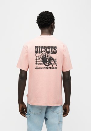 Mann iført lys rosa T-skjorte med korte ermer med svart okse og "Dickies Genuine Workwear"-trykk på baksiden, kombinert med lyseblå jeans.