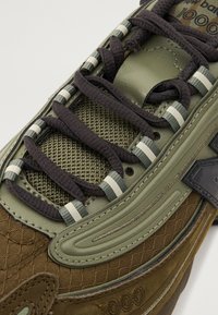 Gros plan sur une basket vert olive et marron avec des lacets noirs, un textile en mesh, et "New Balance 1000" visible sur la languette et le côté.