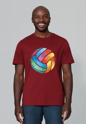 Lachende man die een rood T-shirt draagt met een kleurrijk abstract volleybalontwerp, staand tegen een effen lichtgrijze achtergrond.