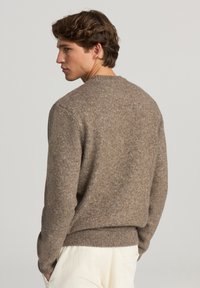 Maglione marrone a maglia con scollatura rotonda, polsini e orlo a coste. Tessuto leggermente testurizzato, indossato con pantaloni color crema.