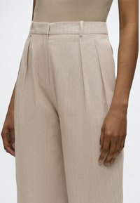 Pantalon beige taille haute, jambes larges, avec fines rayures verticales et plis à l'avant, porté par une personne à la peau marron moyenne.