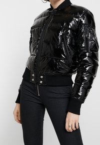 Svart, glansig puffjacka med en figurnära design, ribbade ärmslut och fåll, dragkedjefickor och central dragkedjestängning.