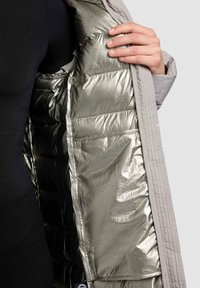 Light gray outer jacket with a metallic silver lining, featuring quilted padding and a pocket inside, held open by a hand.