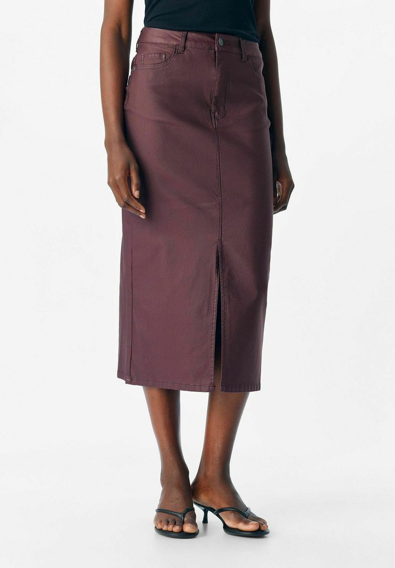 Femme portant une jupe midi violette foncée avec une fente à l'avant et des sandales à talons noires, se tenant devant un fond clair.