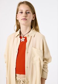 Beige button-up shirt with rolled sleeves, worn over a red ribbed crop top. Necklace features a white flower pendant, adding detail.
