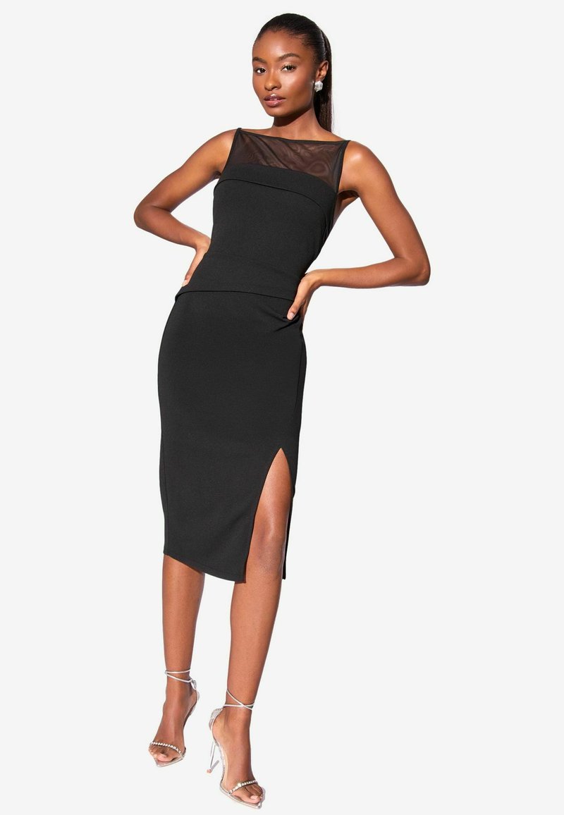 Schwarzes, tailliertes Kleid mit einem transparenten Netz-Ausschnitt, knielang mit einem Seitenschlitz, kombiniert mit klaren Riemchenabsätzen. Glatte Stoffoberfläche und strukturierte Gestaltung.
