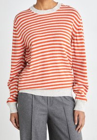 Maglione a righe in arancione e crema, con scollo rotondo e maniche lunghe, abbinato a pantaloni grigi plissettati.