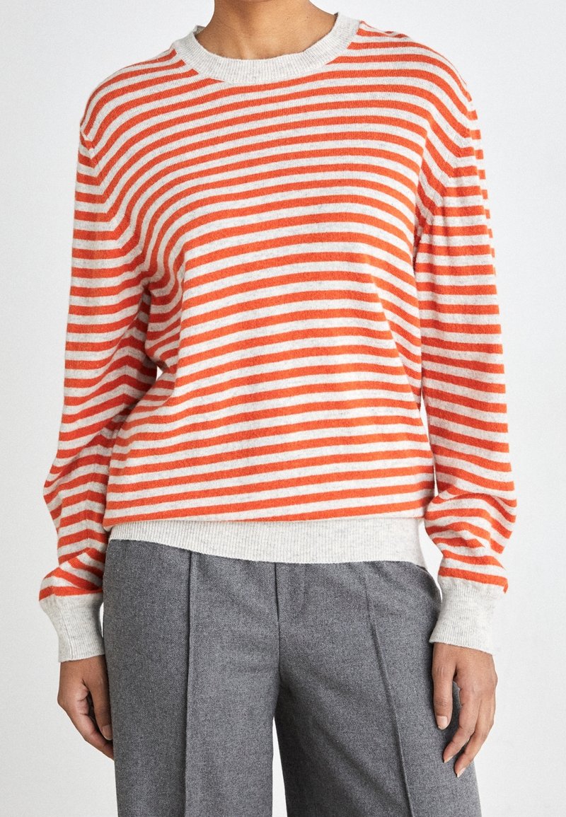 Maglione a righe in arancione e crema, con scollo rotondo e maniche lunghe, abbinato a pantaloni grigi plissettati.