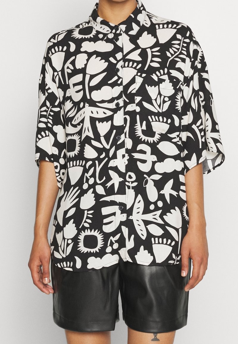 Svart och vit skjorta med kort ärm, försedda med blomster- och abstrakt mönster, i avslappnad passform. Tillsammans med svarta lädershorts.