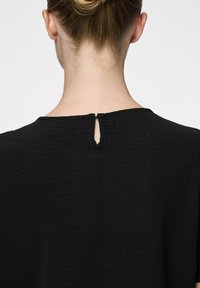 Top nero testurizzato con scollatura rotonda e una piccola apertura a forma di chiave sul retro. Il tessuto appare leggero e leggermente stropicciato.