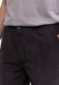 Hombre vistiendo pantalones de pana negros con cierre de botones y camiseta gris claro metida por dentro, con la mano en el bolsillo del lado derecho.