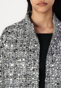 Veste à sequins argentés et noirs avec un tissu texturé, dotée d'un col châle et d'une coupe près du corps. Accentuée par des détails brillants et réfléchissants.