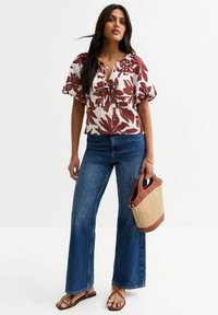 Blouse florale blanche et bordeaux, manches courtes bouffantes, liens à l'avant ; associée à un jean évasé taille haute et un sac en osier marron.