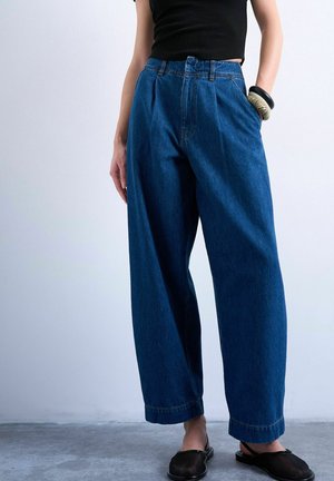 Vrouw draagt blauwe jeans met hoge taille en brede pijpen, zwart mouwloos topje, zwarte instapschoenen en chunky armbanden aan haar linkerpols.