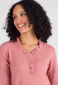 Maglione rosa con scollo a V, dettagli in volant lungo il colletto e tre bottoni decorativi sul torso. Tessuto morbido e testurizzato.