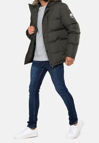 Dunkelgrüne Pufferjacke mit einem fellgefütterten Kapuze, mit Reißverschluss und Knopfverschluss. Getragen über einem grauen Sweatshirt, kombiniert mit blauen Jeans und weißen Turnschuhen.
