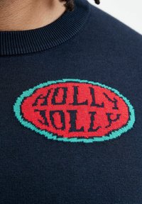 Navy blue knitted sweater featuring a large red oval patch with the text "JOLLY" in black, bordered by a teal accent.