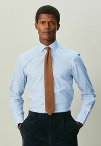 Svetlomodrá károvaná košeľa s štruktúrovaným golierom, doplnená o štruktúrovanú hnedú kravatu. Model má na sebe tmavé nohavice a ruky v vreckách.