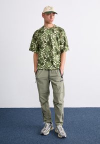 Zeleni s kratkimi rokavi s potiskom, kombiniran z svetlo zelenimi najlon hlačami. Bež kapa z zelenimi poudarki. Siva in bela športna obutev.