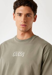 Olivově zelené bavlněné tričko s krátkými rukávy, kulatým výstřihem a bílým grafickým nápisem "GUESS", s nápisem "CLASSIC STYLE & DESIGN" nad ním.