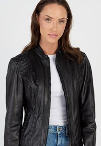 Zwarte leren jas met een ritssluiting aan de voorkant, geribbelde schouderdetails en een soepele textuur. Draagt over een witte top en gecombineerd met blauwe jeans.
