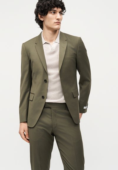 Νέος άνδρας με σγουρά μαλλιά, ντυμένος με ελιά-πράσινο κοστούμι και μπεζ πλεκτή πόλο πουκάμισο, κοιτάζει πλάγια σε ένα απλό φόντο.