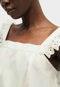 Κοντινό πλάνο ενός ατόμου που φορά λευκό τοπ με τετράγωνη λαιμόκοψη και ριγέ, δαντελωτά μανίκια με οπές στους γυμνούς ώμους.