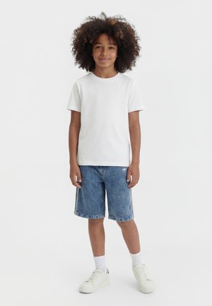 Barn med krøllet hår iført en hvid kortærmet t-shirt, blå denimshorts med sidelommer, hvide sokker og hvide sneakers, der står og smiler.
