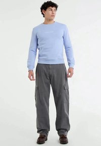 Sweatshirt azul claro com decote redondo e logótipo branco, combinado com calças cargo cinzas com múltiplos bolsos e um corte largo. Sapatos castanhos.