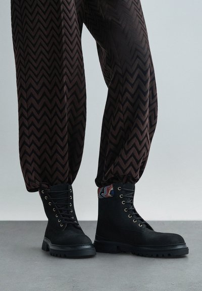 Μαύρες σουέτ πολεμικές μπότες με ανθεκτικούς πάτους, με χρυσά μάτια και μοτίβο υφασμάτινης λεπτομέρειας στη μεριά, συνδυασμένες με καφέ παντελόνι με σχέδια.