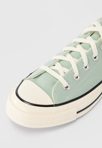 Ανοιχτό πράσινο πάνινο αθλητικό παπούτσι με κρέμ κορδόνια, λαστιχένιο προστατευτικό στη μύτη και μαύρες διακοσμητικές ρίγες. Ασημένιες οπές για λεπτομέρεια κορδονιού.